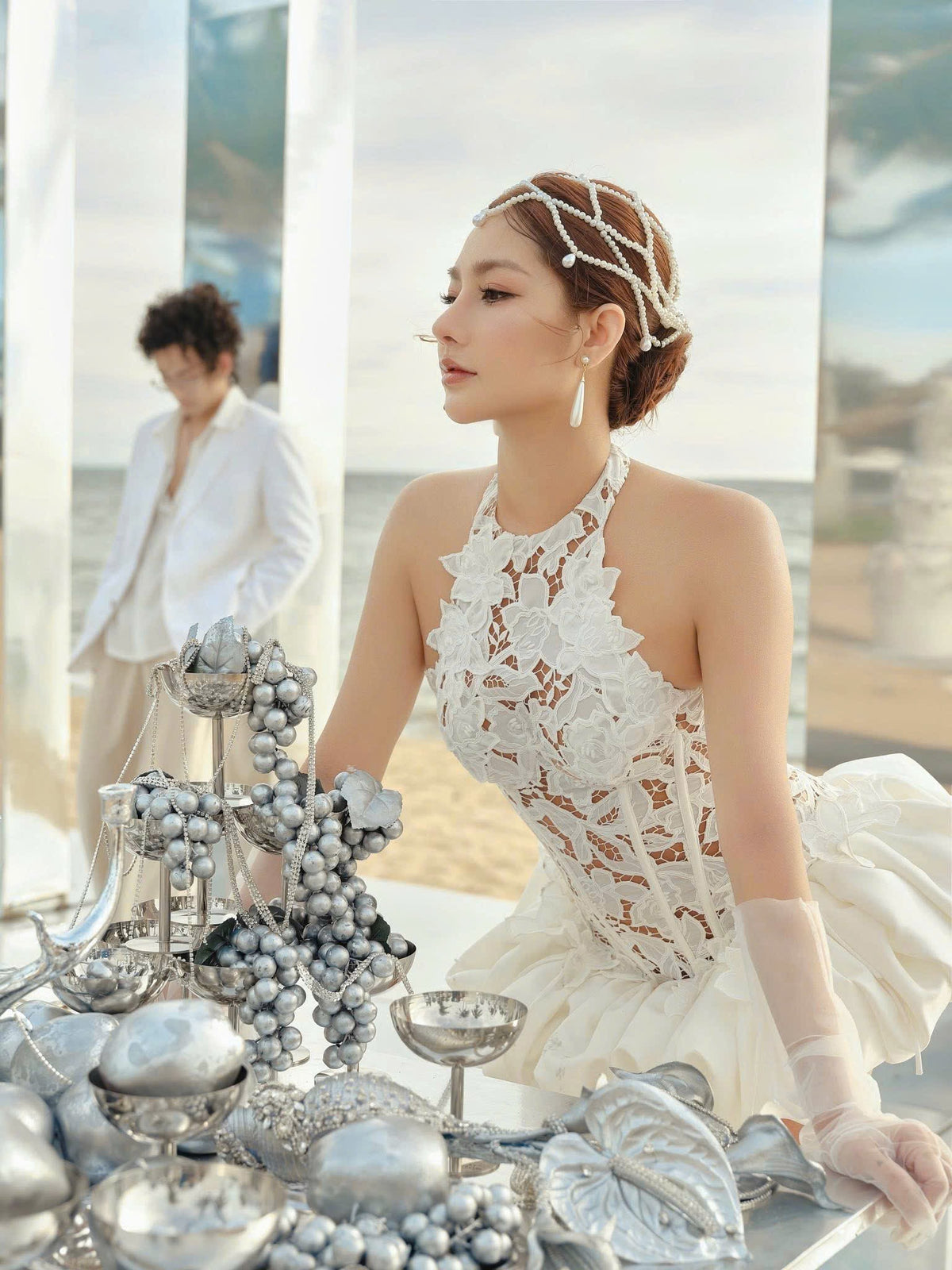 Woman in a white lace dress sitting at a table with silver decorations, blurred background